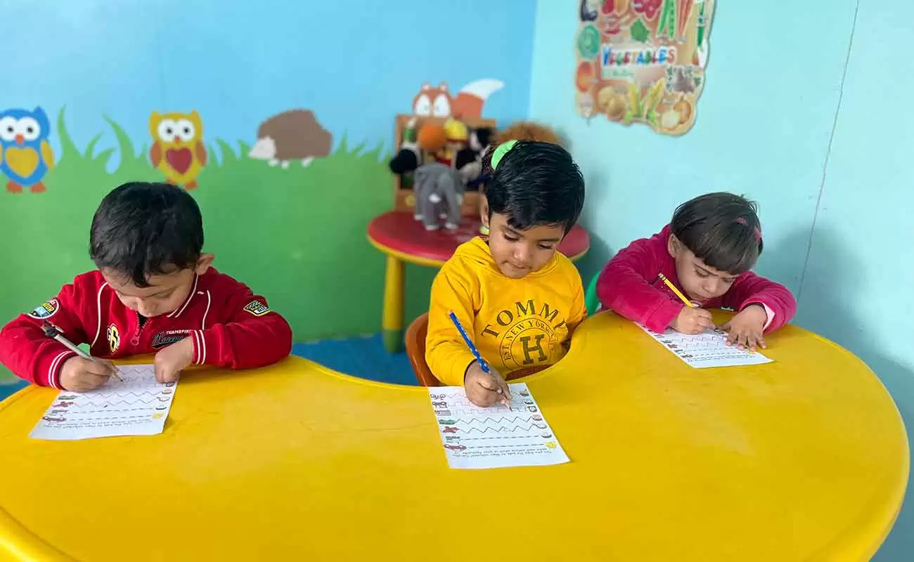 Three young children sitting at a yellow table in a classroom, practicing writing on worksheets with pencils, in a colorful learning environment with playful wall illustrations