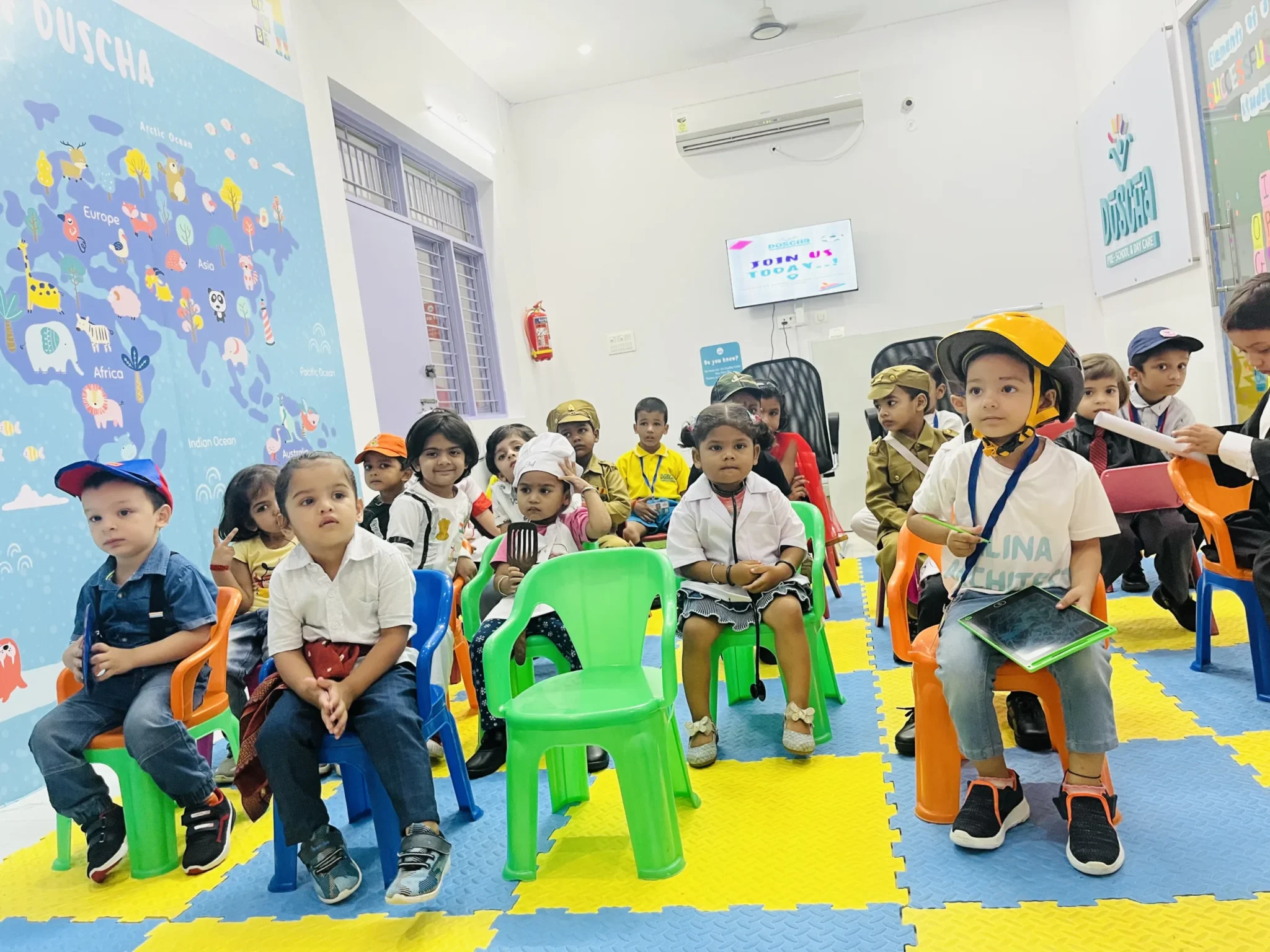 Group of young children attending an activity session at Duscha Preschool and Daycare in Ratan Khand, Lucknow. The children are seated on colorful chairs, dressed in various costumes, including helmets and uniforms, indicating a themed or role-playing event.