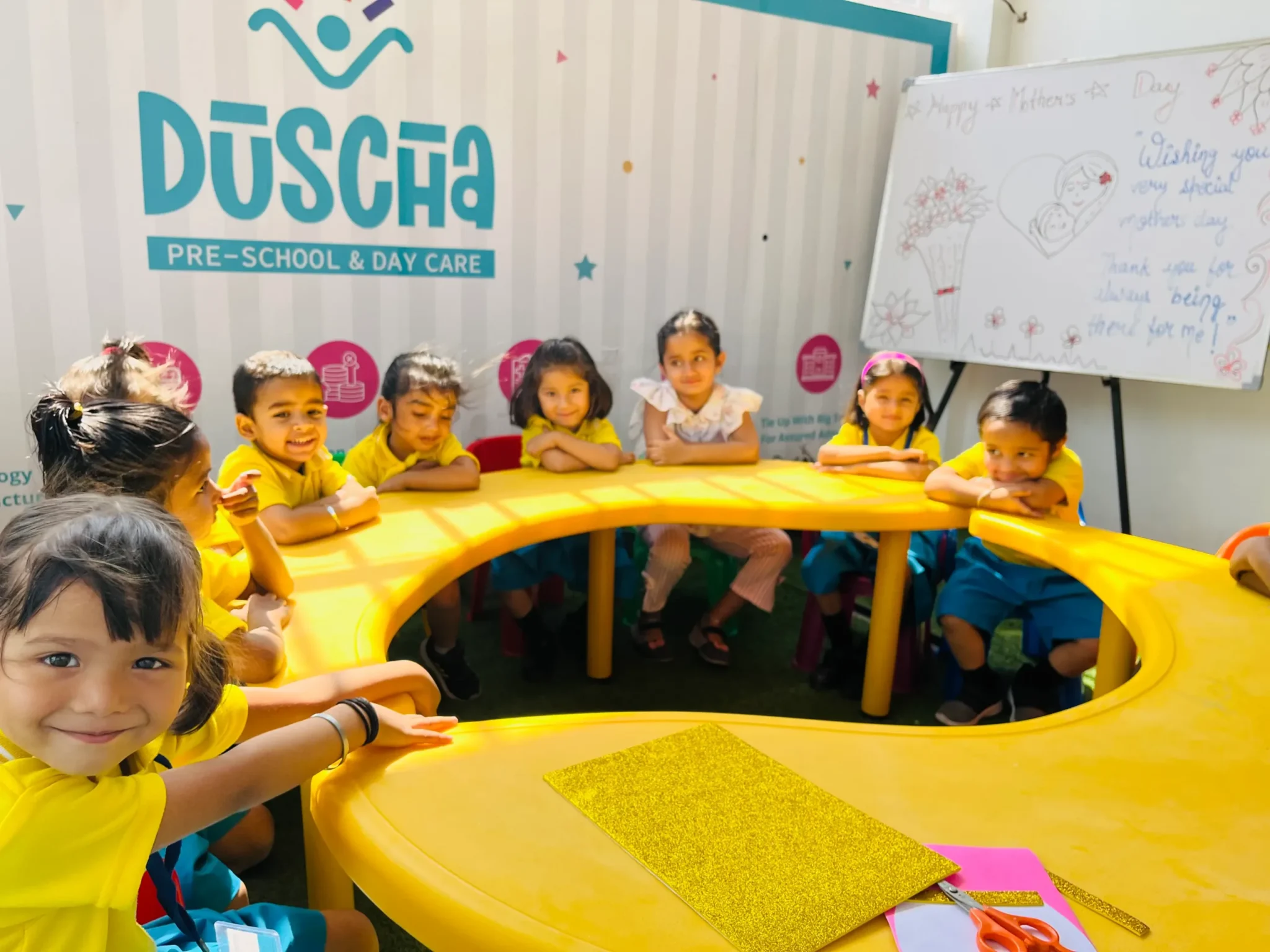 Group of young children sitting around a yellow, curved table in a preschool classroom, smiling and engaged. The classroom has colorful decorations with the name "Duscha Pre-School & Day Care" on the wall in the background.