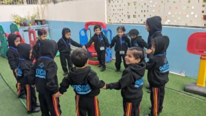 A group of young children dressed in black hoodies, smiling and holding hands in a circle during an outdoor activity, fostering teamwork and social skills through positive parenting practices in a preschool setting.