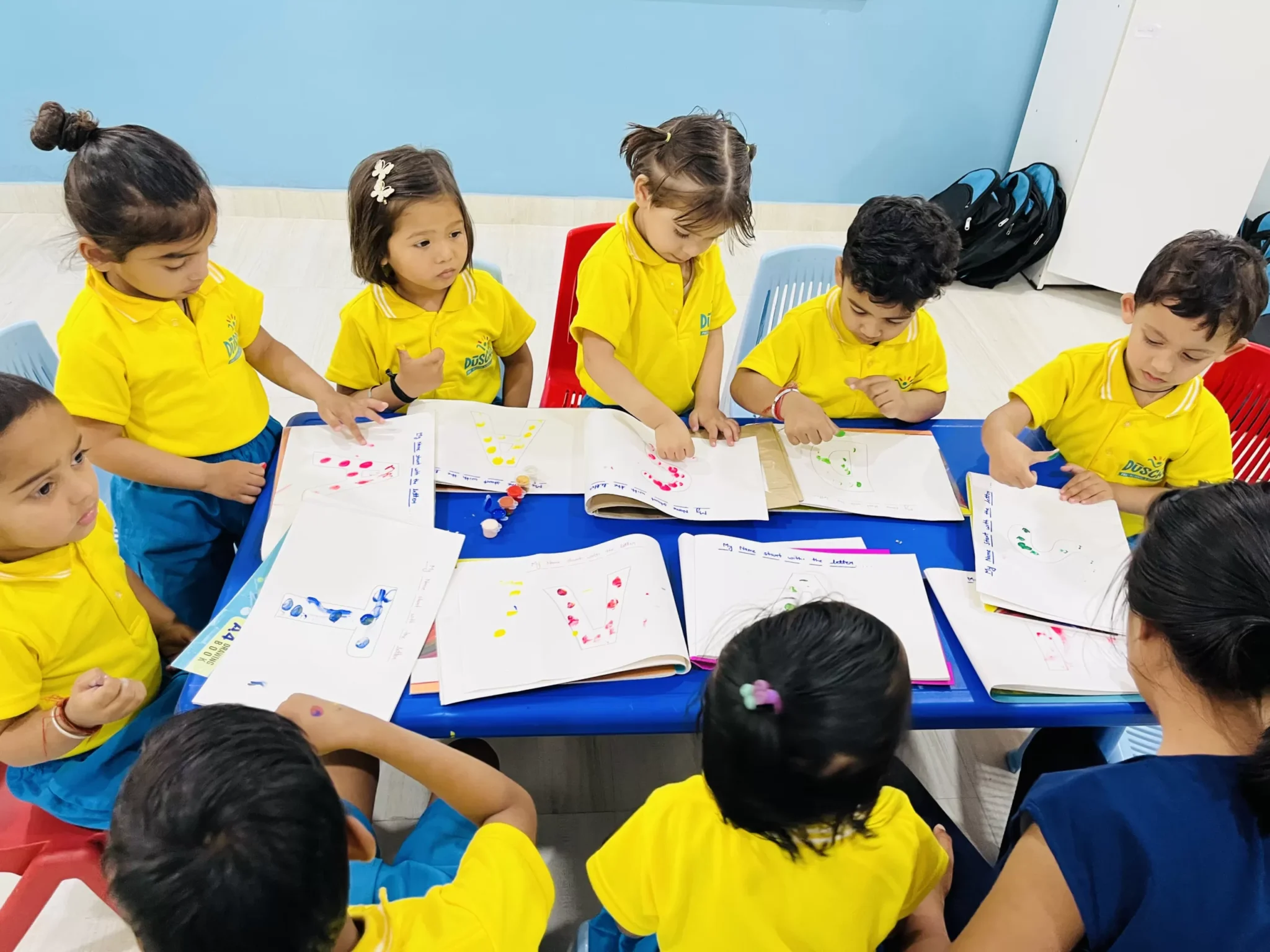 A group of young children in yellow uniforms engaged in a life skills activity, creating colorful art in workbooks at a classroom table, guided by a teacher.