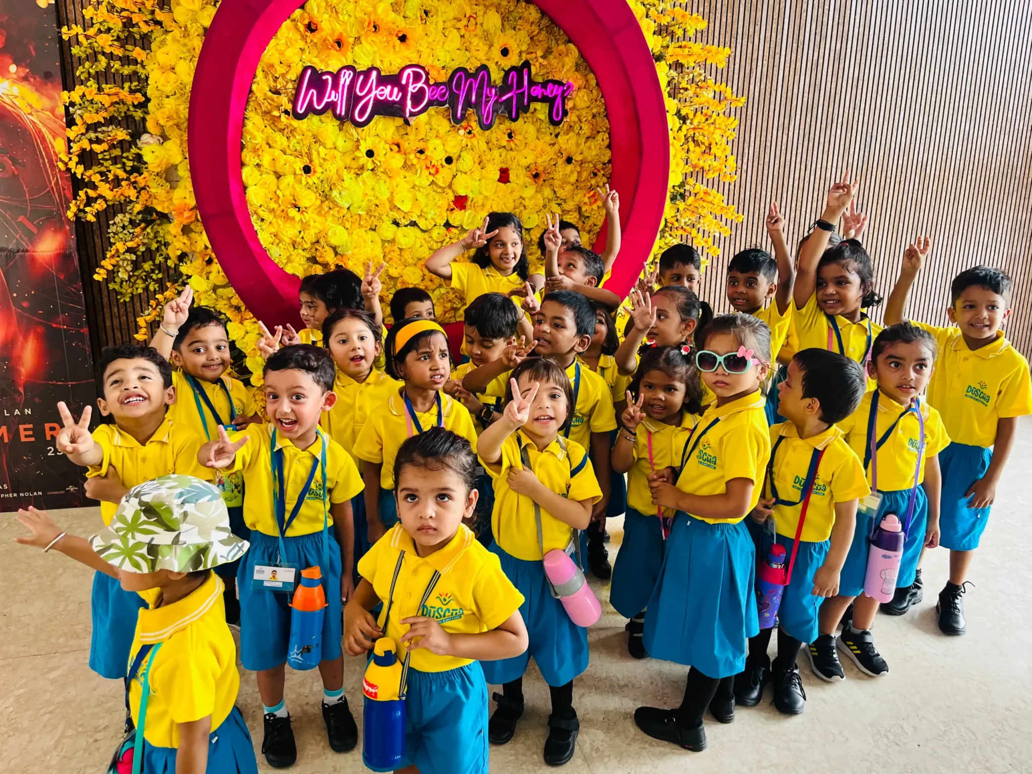 Children at a daycare center in Rajani Khand, learning digital skills in a vibrant and engaging environment, posing happily in their uniforms in front of a colorful floral wall with a neon sign.