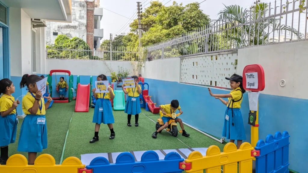 Children in school uniforms participating in a traffic awareness activity in a playground, holding pictures of vehicles and interacting with a child acting as a traffic controller next to a "Traffic Signal" sign.